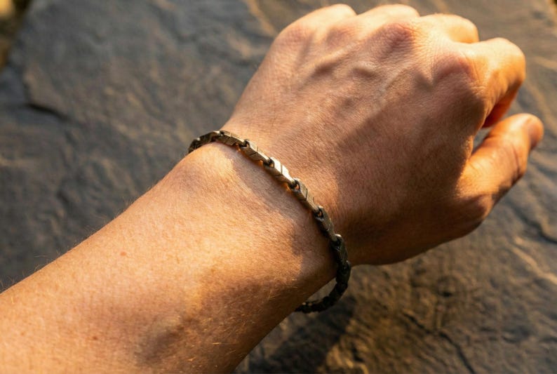May include: A silver-toned bracelet with a geometric design is worn on a wrist. The bracelet is composed of interconnected, arrow-shaped links. The skin tone is light, and the background is a blurred, natural setting.