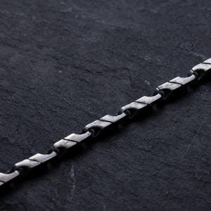 May include: A silver-toned chain necklace with a unique, geometric design. The chain is composed of interconnected, stylized links, creating a textured appearance. The necklace is displayed against a dark, textured background.