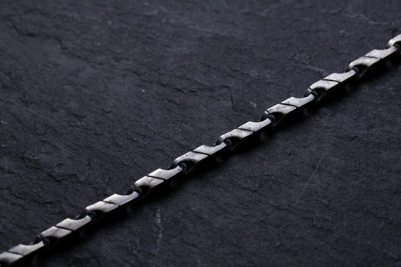 May include: A silver-toned chain necklace with a unique, geometric design. The chain is composed of interconnected, stylized links, creating a textured appearance. The necklace is displayed against a dark, textured background.