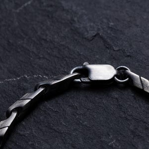 May include: Close-up of a silver-toned bracelet clasp and chain. The chain has a geometric design with a textured, oxidized finish. The clasp is securely fastened, showcasing the bracelet's construction against a dark, textured background.
