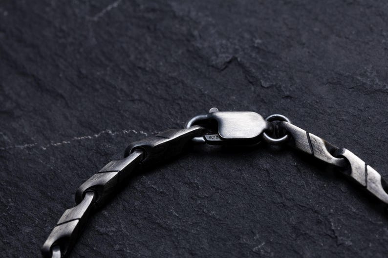 May include: Close-up of a silver-toned bracelet clasp and chain. The chain has a geometric design with a textured, oxidized finish. The clasp is securely fastened, showcasing the bracelet's construction against a dark, textured background.