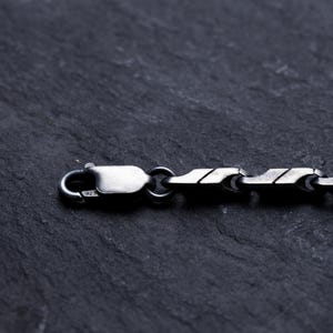May include: Close-up of a silver chain necklace with a unique, geometric link design. The necklace features a lobster clasp closure. The jewelry is displayed on a dark, textured surface.