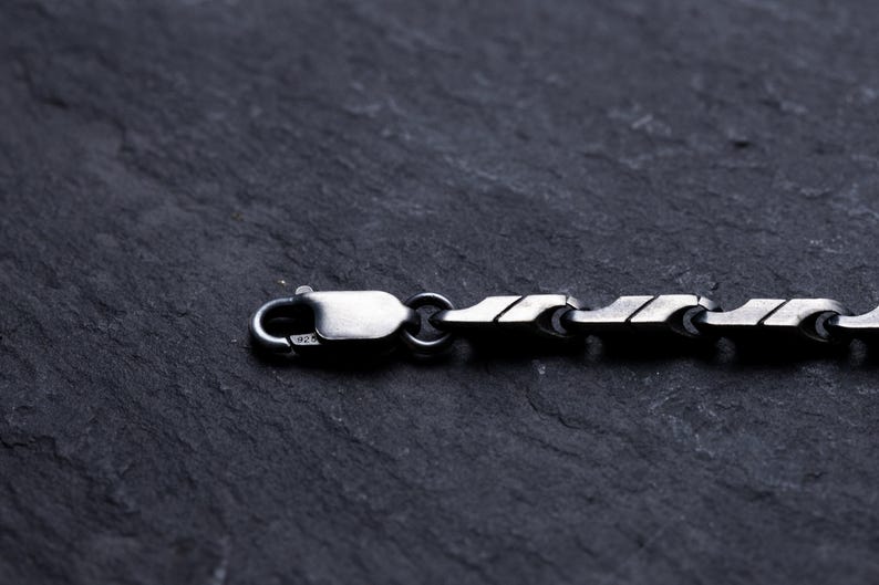 May include: Close-up of a silver chain necklace with a unique, geometric link design. The necklace features a lobster clasp closure. The jewelry is displayed on a dark, textured surface.