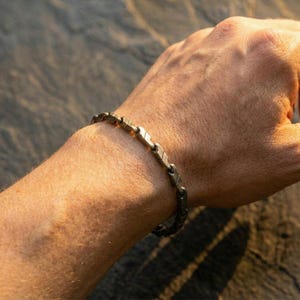 May include: A silver-toned bracelet with a linked design is worn on a wrist. The bracelet's links have a textured appearance. The image is lit with warm sunlight, highlighting the details of the jewelry.