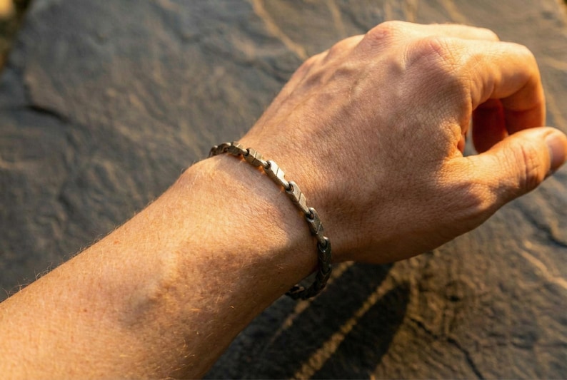 May include: A silver-toned bracelet with a linked design is worn on a wrist. The bracelet's links have a textured appearance. The image is lit with warm sunlight, highlighting the details of the jewelry.