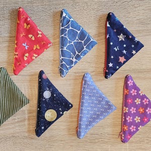 Könnte beinhalten: Eine Sammlung dreieckiger Stoffbeutel mit Reißverschlüssen, jeweils mit einem einzigartigen Muster. Designs umfassen Schmetterlinge, blauen Giraffenmuster, Sterne, Streifen, Planeten, Anker und Blumen. Die Beutel sind auf einer hellen Holzoberfläche angeordnet.