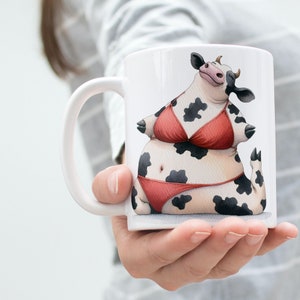 May include: White ceramic mug with a black and white cow wearing a red bikini. The cow is sitting on a white background.