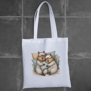 May include: White tote bag with a graphic of two capybaras wearing sleep masks and pajamas, cuddling in bed.