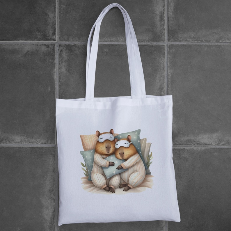 May include: White tote bag with a graphic of two capybaras wearing sleep masks and pajamas, cuddling in bed.