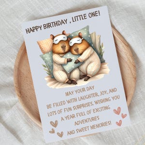 May include: A birthday card with a light blue background featuring two capybaras wearing pajamas and sleeping with a pillow. The card has the text "Happy Birthday, Little One!" and a message wishing a year full of exciting adventures and sweet memories.