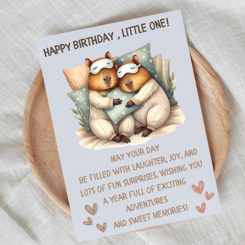 May include: A birthday card with a light blue background featuring two capybaras wearing pajamas and sleeping with a pillow. The card has the text "Happy Birthday, Little One!" and a message wishing a year full of exciting adventures and sweet memories.