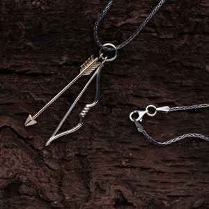 Op de afbeelding: Een zilveren ketting met een zilveren boog en pijl hanger. De pijl is goud. De ketting ligt op een bruine houten achtergrond.
