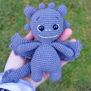 Puede incluir: Un juguete de dragón de crochet gris con ojos negros y una sonrisa blanca. El dragón tiene alas y una cola.