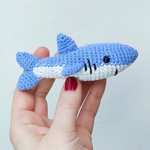 Peut inclure: Un jouet en crochet en forme de requin bleu et blanc avec des yeux noirs. Le requin est tenu dans une main.