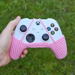 May include: A white Xbox controller with pink crocheted covers on the handles. The controller has the standard buttons: A, B, X, Y, and a directional pad.