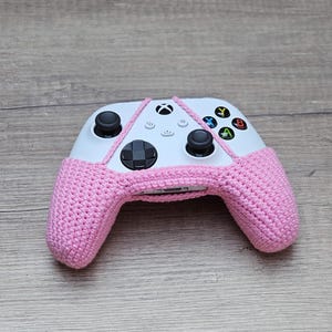 May include: A pink crocheted cover for a white Xbox controller. The controller has the standard Xbox buttons, including the A, B, X, and Y buttons.