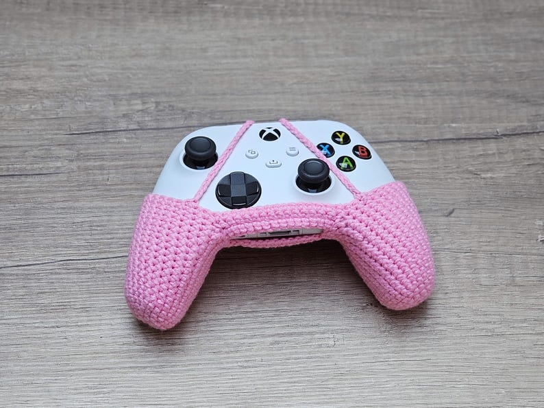 May include: A pink crocheted cover for a white Xbox controller. The controller has the standard Xbox buttons, including the A, B, X, and Y buttons.