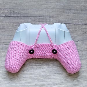 May include: A pink crocheted cover for a video game controller. The cover has two black buttons on the front.