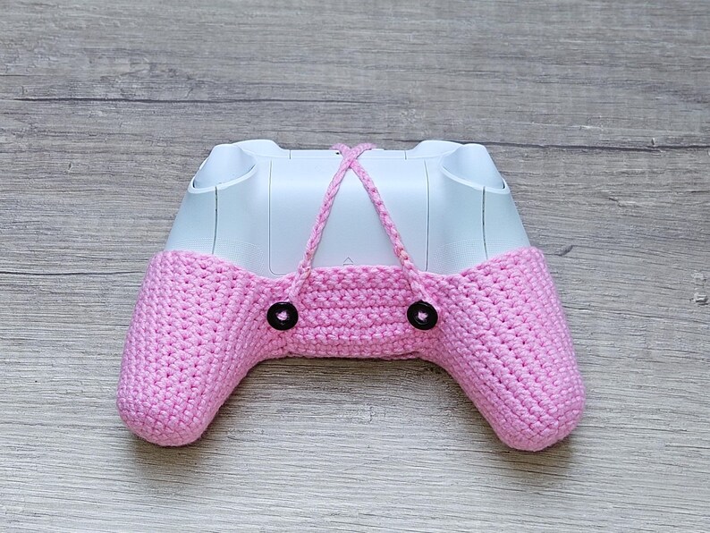 May include: A pink crocheted cover for a video game controller. The cover has two black buttons on the front.