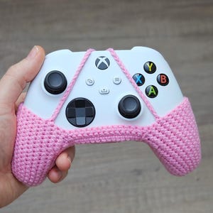 May include: A white Xbox controller with pink crocheted covers over the handles. The controller has the standard buttons: A, B, X, Y, and a directional pad.