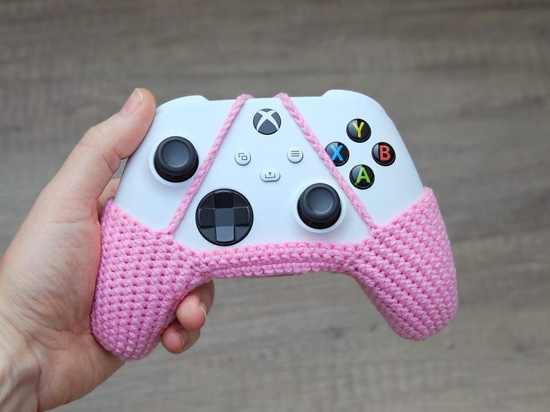 May include: A white Xbox controller with pink crocheted covers over the handles. The controller has the standard buttons: A, B, X, Y, and a directional pad.