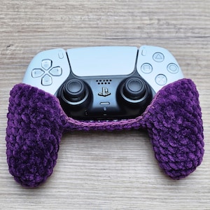 May include: A black and white video game controller partially covered by a deep purple, textured, hand-crocheted yarn sleeve. The controller features a central PlayStation logo, two black analog sticks, a directional pad, and four action buttons.