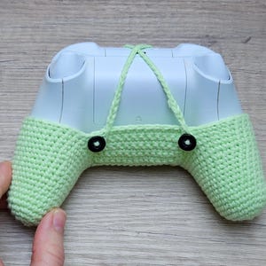 May include: A light green crocheted cover for a video game controller. The cover has two black buttons on the back and a white controller is visible inside.