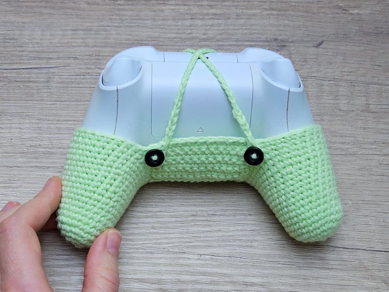 May include: A light green crocheted cover for a video game controller. The cover has two black buttons on the back and a white controller is visible inside.