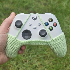 May include: A white Xbox controller with a light green crocheted cover. The cover has a strap that goes around the controller and ties at the top.