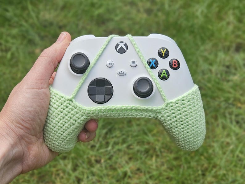May include: A white Xbox controller with a light green crocheted cover. The cover has a strap that goes around the controller and ties at the top.