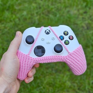 Puede incluir: Un mando Xbox blanco con cubiertas de crochet rosa. El mando tiene un diseño blanco y negro con botones etiquetados como X, Y, A y B.