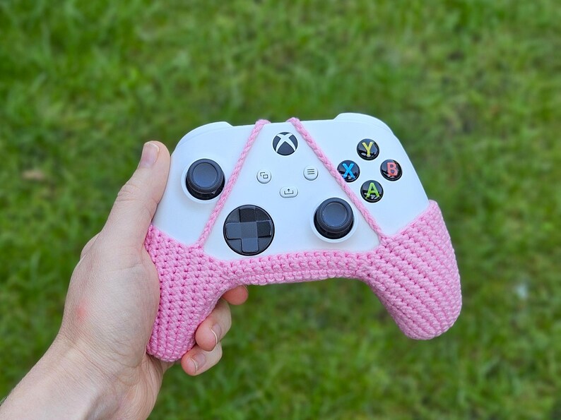May include: A white Xbox controller with pink crocheted covers. The controller has a white and black design with buttons labeled X, Y, A, and B.