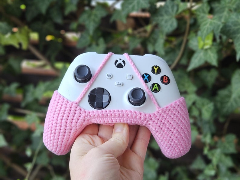 May include: A white Xbox controller with pink crocheted covers over the handles. The controller has the standard Xbox buttons, including A, B, X, and Y.