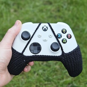 May include: A white Xbox controller with black crocheted covers on the handles. The controller has a black D-pad, black buttons, and a white Xbox logo.