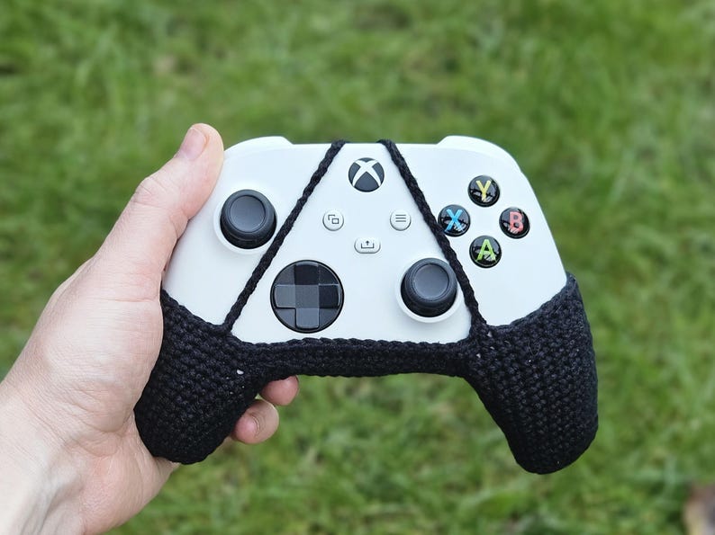 May include: A white Xbox controller with black crocheted covers on the handles. The controller has a black D-pad, black buttons, and a white Xbox logo.