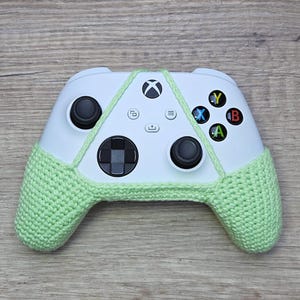 May include: A white Xbox controller with a light green crocheted cover. The controller has the standard buttons, including A, B, X, and Y.