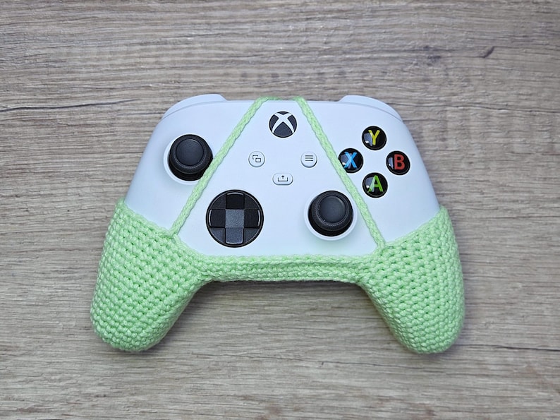 May include: A white Xbox controller with a light green crocheted cover. The controller has the standard buttons, including A, B, X, and Y.
