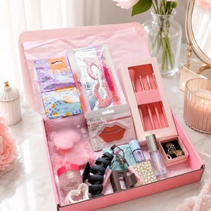Op de afbeelding: Een roze geschenkdoos met diverse beauty-artikelen, waaronder een set make-upkwasten, collageen lippads, oogmaskers, een metalen wimperkruller, haarklemmen, een potje met glitters, een flesje nagellak en een kleine goudkleurige ketting.