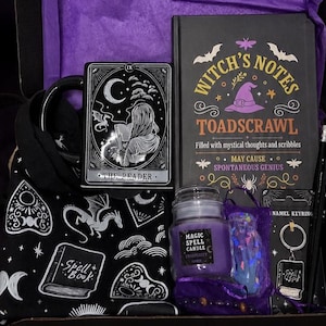 Könnte beinhalten: Eine Geschenkbox mit einer schwarzen Tasse mit Tarotkarten-Design, einem schwarzen Notizbuch mit dem Titel Witch's Notes Toadscrawl, einer lila Kerze mit der Aufschrift Magic Spell Candle, einem schwarzen Emaille-Schlüsselanhänger, zwei schwarzen Stiften.