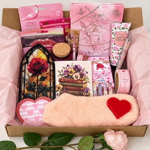 Puede incluir: Una caja de regalo llena de artículos de color rosa que incluye un diario con estampado de rosas, crema de manos, mascarillas de colágeno, una vela, un adorno de rosa estilo vidriera, una tarjeta floral, cinta adhesiva, un bolígrafo y calcetines suaves con un corazón rojo.