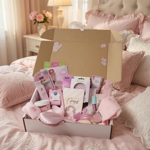 May include: A cardboard gift box filled with various pink-themed beauty and self-care items, including a satin eye mask, a small jar, a hair brush, a pink headband, a lip gloss, and a card with the text "AMS Free" printed in gold script.