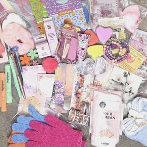 Op de afbeelding: Een verzameling schoonheids- en kantoorartikelen, waaronder roze scrunchies, markeerstiften, pennen, een gua sha-tool, gezichtsmaskers, hartvormige accessoires, kralenarmbanden, bloemrijke bladwijzers, washi-tape, scrubhandschoenen en een haartulband.