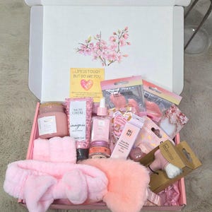 Könnte beinhalten: Eine rosa Geschenkbox gefüllt mit Schönheits- und Pflegeprodukten. Die Box enthält ein rosa Stirnband, eine Augenmaske, Hautpflegeprodukte und eine Kerze. Der Deckel der Box zeigt ein Blumendesign und den Text "LIFE IS TOUGH BUT SO ARE YOU."