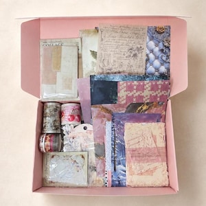 May include: A light pink cardboard box filled with various stationery supplies, including rolls of patterned washi tape, assorted decorative paper sheets with vintage-style prints, floral illustrations, and small cards with text and graphic designs.