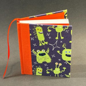 Puede incluir: Un pequeño cuaderno hecho a mano con un vibrante estampado de monstruos en la portada. La cubierta presenta monstruos verde lima sobre un fondo azul oscuro, acentuados con estrellas blancas. El lomo es de color naranja brillante y una cinta roja sirve de marcador.