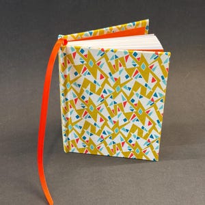 Puede incluir: Un diario de tapa dura con un patrón geométrico en amarillo, blanco, rojo y azul. El diario tiene una cinta naranja como marcador. Las páginas son blancas y las cubiertas interiores son naranjas.