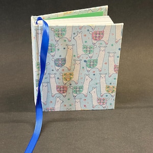 Puede incluir: Un pequeño cuaderno de tapa dura con una cubierta azul claro que presenta un patrón repetido de llamas blancas con coloridas mantas de silla y pequeñas flores. Una cinta azul brillante como marcador se extiende desde la parte superior.