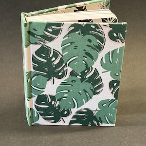 Puede incluir: Un cuaderno de tapa dura con un estampado de hojas de monstera verde y blanco. El cuaderno está abierto, revelando las páginas blancas en el interior. El lomo y los bordes son verdes, y la cubierta presenta hojas grandes y estilizadas.