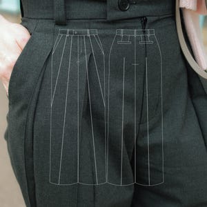 May include: Close-up of dark grey pleated trousers with a button closure and belt loops. The trousers have a high waist and a tailored fit. A hand is in the pocket. The image also shows a technical drawing of the trousers.
