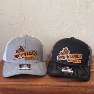 May include: Two trucker hats with a brown leather patch that says "DROP'N LOADS & FILL'N HOLES" with a dump truck graphic. One hat is gray with a white mesh back and the other is black with a gray mesh back. Both hats have a Richardson 112 label.